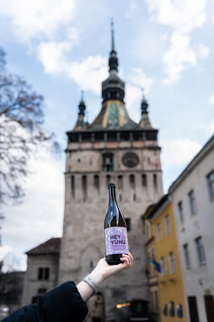 A hand raising a bottle of Voyage Kombucha in front of the Sighișoara clock tower