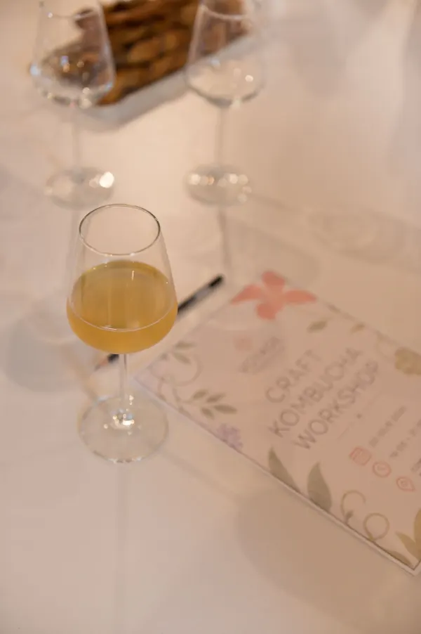 A glass of kombucha next to the printed Craft Kombucha Workshop guide on a white table